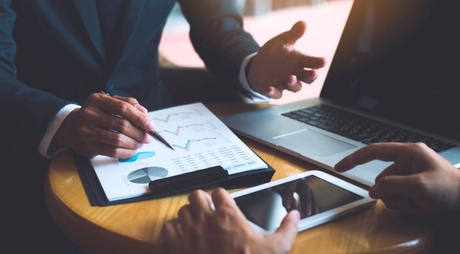 Two businessmen are discussing and analyzing the performance of the company business paper and looking for numerical data via tablets.